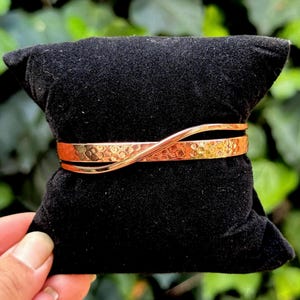 Può includere: Un bracciale color rame con una texture martellata e un design attorcigliato. Il bracciale è esposto su un cuscino di velluto nero, con fogliame verde sullo sfondo. Un accessorio elegante.