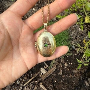 May include: Gold-toned oval locket necklace with a starburst design and a green gemstone center. The locket hangs from a delicate chain. The necklace is held in a hand against a backdrop of soil and greenery.
