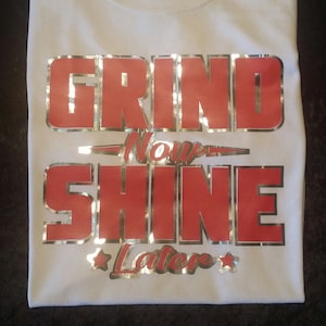Può includere: Maglietta bianca con il testo "GRIND Now SHINE Later" in lettere rosse e argentate.