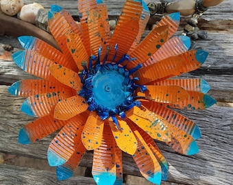May include: A large, orange and blue metal flower with a textured surface. The flower has a blue centre and is made from recycled materials.