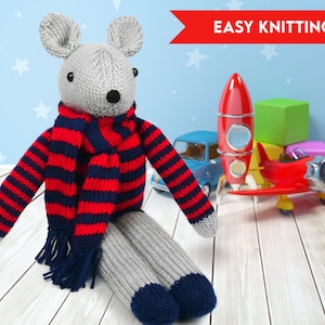 May include: A grey knitted mouse wearing a red and blue striped jumper and scarf. The mouse is sitting on a wooden surface with toy aeroplanes and a rocket ship in the background. The text "EASY KNITTING" is in red at the top of the image.