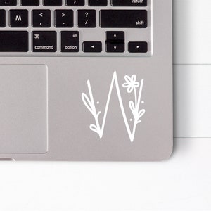 May include: A white laptop with a floral design sticker on the trackpad. The sticker features a stylized mountain range with a flower and leaves.