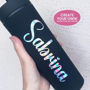 May include: A matte black cylindrical tumbler featuring the name Sabrina in iridescent, holographic script lettering with a small heart icon. A pink oval graphic in the upper right corner contains the text CREATE YOUR OWN! CHOOSE COLOR FONT AND SIZE!