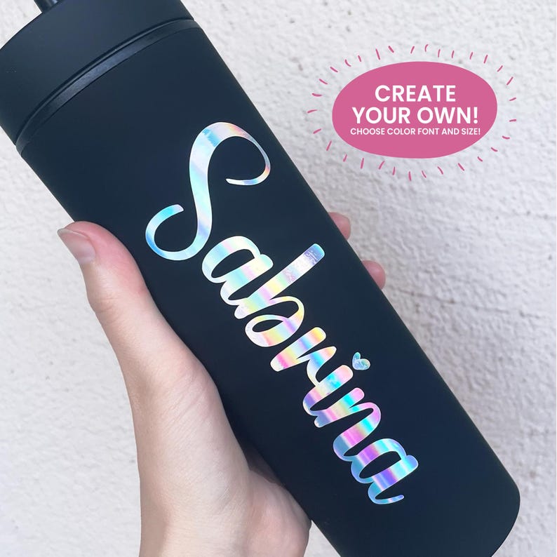 May include: A matte black cylindrical tumbler featuring the name Sabrina in iridescent, holographic script lettering with a small heart icon. A pink oval graphic in the upper right corner contains the text CREATE YOUR OWN! CHOOSE COLOR FONT AND SIZE!