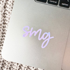May include: A holographic sticker with the letters "smg" in a cursive font. The sticker is a light purple color and is placed on a silver laptop.