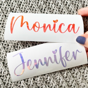 May include: Two custom vinyl decals on white backing paper. The top decal features the name Monica in a gradient orange-to-red script font with a small heart above the i. The bottom decal displays the name Jennifer in a holographic script font.