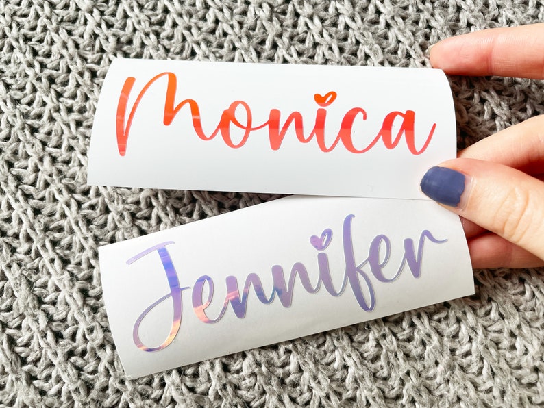 May include: Two custom vinyl decals on white backing paper. The top decal features the name Monica in a gradient orange-to-red script font with a small heart above the i. The bottom decal displays the name Jennifer in a holographic script font.
