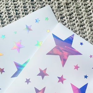 K&ouml;nnte beinhalten: Zwei Bl&auml;tter wei&szlig;es Papier mit schimmernden, holografischen Stern-Aufklebern. Die Sterne sind in verschiedenen Gr&ouml;&szlig;en und haben einen Regenbogeneffekt.