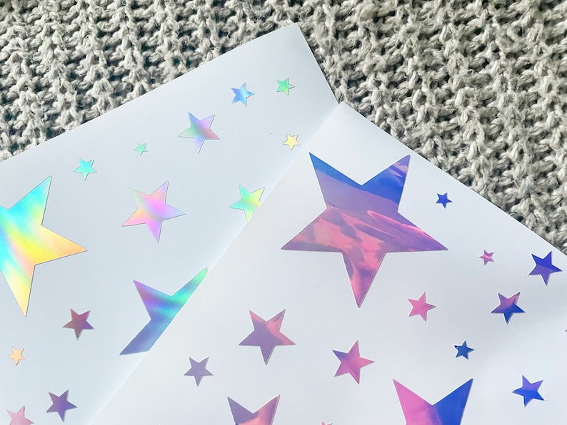 K&ouml;nnte beinhalten: Zwei Bl&auml;tter wei&szlig;es Papier mit schimmernden, holografischen Stern-Aufklebern. Die Sterne sind in verschiedenen Gr&ouml;&szlig;en und haben einen Regenbogeneffekt.