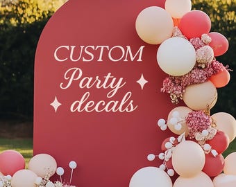 以下が含まれることがあります： 「CUSTOM Party decals」という白い文字と四芒星のアクセントが施された、大きなアーチ型のマットな赤色のディスプレイボード。ボードは、クリーム色、白、ピンクの色合いのバルーンの房と、ドライフラワーで装飾されています。