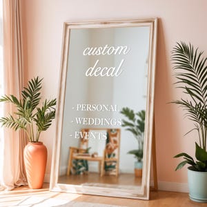 May include: A large, rectangular mirror with a distressed white frame leans against a wall. The mirror reflects a room with plants and furniture. Text on the mirror reads "custom decal" followed by "- PERSONAL", "- WEDDINGS", and "- EVENTS".