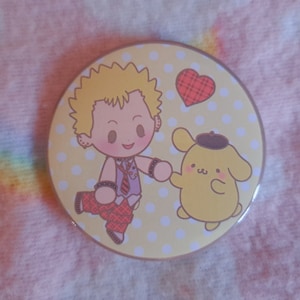 May include: A circular badge with a cartoon illustration on a yellow background with white polka dots. The design features a character with blonde hair and a dog-like character holding hands, with a red heart.