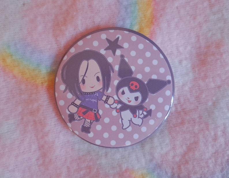 May include: A circular badge with a light purple background and white polka dots. The badge features two cartoon characters holding hands. One character has black hair and a purple top. The other is black and white with a red skull.