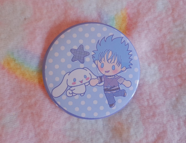 May include: A circular badge with a light blue background and a cartoon illustration. The design features a blue-haired character and a white bunny-like creature holding hands. A plaid star and white polka dots are also included.