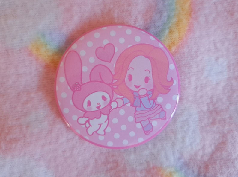 May include: A circular pink badge featuring a cartoon illustration of a girl and a bunny. The badge has a pink polka dot background and a pink border. The girl has red hair and is wearing a blue and pink outfit. The bunny is pink.