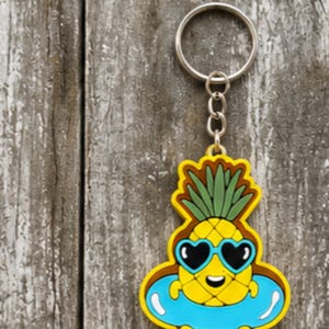 May include: A yellow pineapple keychain featuring a green leafy top, wearing blue heart-shaped sunglasses, and floating in a bright blue inner tube. The pineapple has a small smiling face and is attached to a silver metal split ring by a short chain.