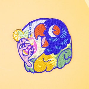 May include: Embroidered patch featuring a colourful parrot holding a tropical drink with a lemon wedge. The parrot is blue, green, yellow, and orange. The drink is pink, yellow, and orange. The lemon wedge is yellow.