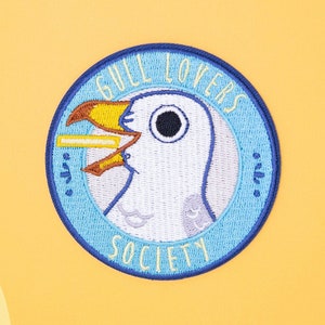 Może przedstawiać: Haftowana naszywka z wizerunkiem kreskówki mewy. Mewa ma żółty dziób i jest umieszczona w niebieskim okręgu. Tekst "GULL LOVERS SOCIETY" jest wygięty wokół okręgu. Tło to żółty gradient.