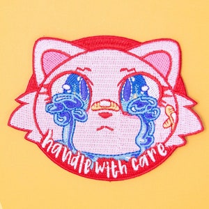 May include: An embroidered patch featuring a cartoon cat face with blue teardrop-shaped eyes. The text "handle with care" is written in a red circle around the cat's face.