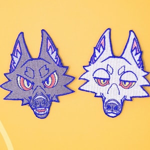 May include: Two embroidered iron-on patches of wolf faces. The wolf on the left is purple with red eyes and a snarling expression. The wolf on the right is white with pink ears and a sleepy expression. Both wolves have blue outlines.
