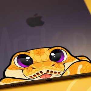 May include: A sticker of a cartoon snake with large purple eyes peeking over the edge of a grey laptop. The snake is yellow and orange with a black tongue sticking out.