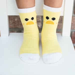 May include: A pair of yellow socks with a white cuff and a cartoon chick face design. The socks are worn on a child's feet.