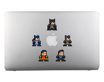 Op de afbeelding: Een zilveren laptop met vijf gepixelde superhelden stickers. De stickers zijn van Batman, Robin en Batgirl. De stickers zijn in een cirkel rond het Apple-logo op de laptop gerangschikt.