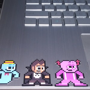 Puede incluir: Tres pegatinas de arte de p&iacute;xeles en un port&aacute;til plateado. Las pegatinas son de un fantasma azul con un sombrero amarillo, un hombre con un traje negro y una criatura rosa con cuernos.