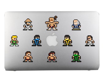 Op de afbeelding: Een zilveren laptop met tien pixel art stickers van personages uit een vechtvideospel. De personages staan in verschillende poses en omvatten een blonde vrouw, een man met een witte baard, een man met een zwart hoofdband, een man met een geel hoofdband, een man met een groen hoofdband, een man met een rood hoofdband, een man met een bruin hoofdband, een man met een rood hoofdband, een man met een bruin hoofdband en een man met een rood hoofdband.
