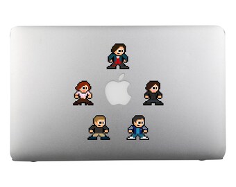 Op de afbeelding: Vijf pixel art stickers van personages uit een tv-programma zijn geplaatst op een zilveren laptop. De personages dragen verschillende gekleurde kleding en staan in verschillende poses.