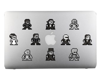 Op de afbeelding: Een zilveren laptop met negen zwart-witte pixel art stickers van klassieke horrorfilmfiguren. De personages omvatten Frankenstein's monster, Dracula, de weerwolf, de mummie, het Wezen uit de Zwarte Lagune, en meer.