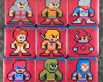 Op de afbeelding: Een set van negen vierkante buttons met pixel art personages uit de Masters of the Universe franchise. Elke button toont een uniek personage op een levendige rode achtergrond met een blauwe rand, in een retro 8-bit grafische stijl.
