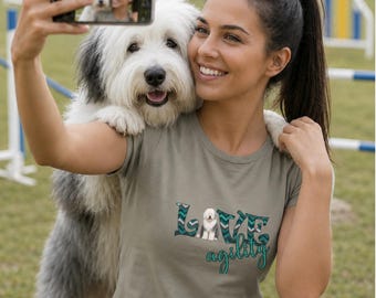 May include: A sage green short-sleeved t-shirt featuring a graphic design with the word LOVE in a teal and grey chevron pattern, with the letter O replaced by an Old English Sheepdog, and the word agility written in teal script below it.
