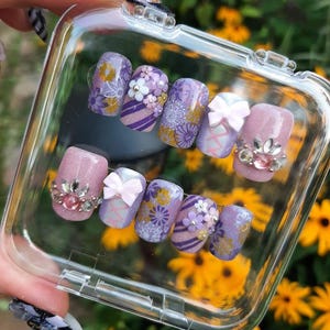 May include: A set of purple and gold nail art tips with floral and geometric designs. The tips are arranged in a clear plastic container.
