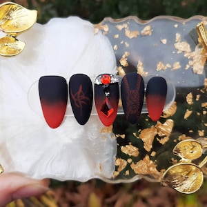 May include: Five black and red ombre press-on nails with different designs. One nail has a red rhinestone accent.