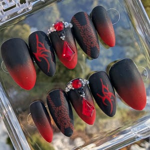 May include: A set of ten almond-shaped press-on nails featuring a matte black and red gradient finish. The design includes red tribal patterns, intricate rope knot illustrations, and central nails adorned with red crystals and clear rhinestones.