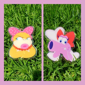 May include: Two colorful cartoon character keychains. The first keychain is yellow with pink and white polka dots and a pink mouth. The second keychain is pink with white and red accents. Both keychains are on a green grassy background.