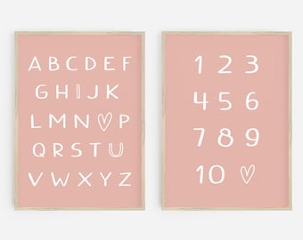 May include: Two framed prints with a light pink background. One print displays the alphabet in white, handwritten-style letters. The other print shows the numbers 1 through 10 in white, with a heart symbol next to the number 10. The frames are a light wood color.