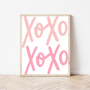 K&ouml;nnte beinhalten: Ein gerahmter Druck mit dem Text "XOXO", der zweimal in Rosa und Hellrosa auf wei&szlig;em Hintergrund wiederholt wird.