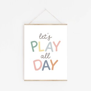 May include: A white framed print with the text "let's PLAY all DAY" in colorful, handwritten letters. The letters are in shades of green, yellow, blue, pink, and orange.