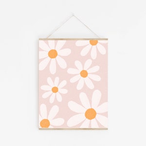 May include: A pink background with a pattern of white daisies with orange centers. The daisies are arranged in a random pattern and are of varying sizes. The print is framed with a light wood frame.