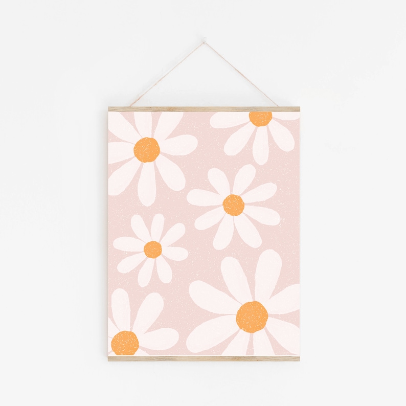 May include: A pink background with a pattern of white daisies with orange centers. The daisies are arranged in a random pattern and are of varying sizes. The print is framed with a light wood frame.