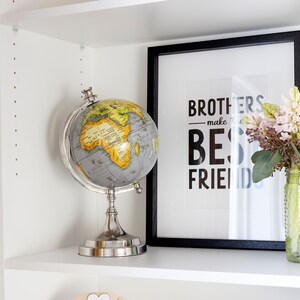 K&ouml;nnte beinhalten: Ein wei&szlig;es B&uuml;cherregal mit einem Globus, einem gerahmten Druck mit dem Text "Brothers make the best friends", einer Vase mit rosa Blumen und einem roten Spielzeuglaster in einem Regal.