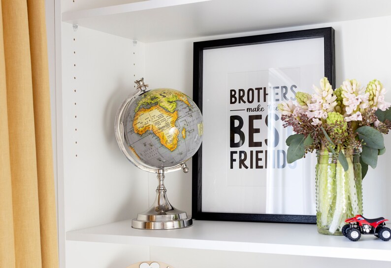 K&ouml;nnte beinhalten: Ein wei&szlig;es B&uuml;cherregal mit einem Globus, einem gerahmten Druck mit dem Text "Brothers make the best friends", einer Vase mit rosa Blumen und einem roten Spielzeuglaster in einem Regal.