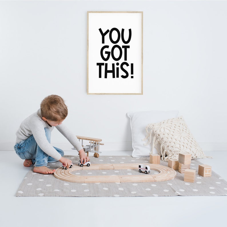 May include: A child plays with a wooden toy race track and toy cars on a grey and white polka dot rug. A framed print with the text "YOU GOT THIS!" hangs on the wall behind the child.