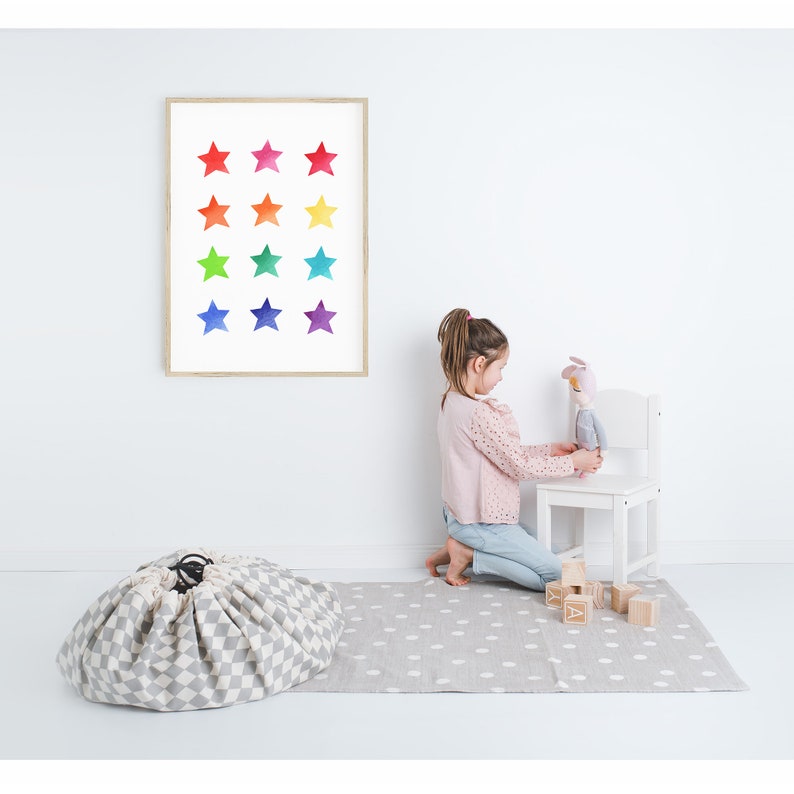 May include: A child plays with a toy on a white chair in a room with a gray and white play mat. A gray and white storage bag is on the floor. A framed print with a rainbow of stars is on the wall.