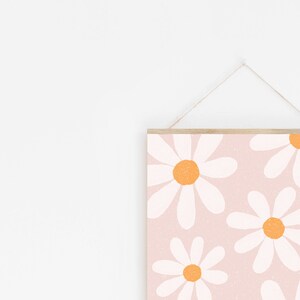 May include: A pink and white print with a pattern of white daisies with orange centers. The print is hanging on a white wall with a wooden frame and twine.