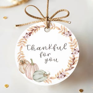 Może przedstawiać: Okrągła etykieta prezentowa z napisem "Thankful for you" w czarnym skrypcie. Etykieta przedstawia akwarelowy wzór dyni i wieniec kwiatowy w odcieniach brązu, różu i zieleni. Jest zawiązana naturalną kokardą ze sznurka.