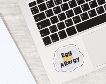 May include: A laptop with a sticker reading "Egg Allergy". The sticker is designed as a fried egg with a yellow yolk and a textured white background. A notebook is visible in the corner of the image.