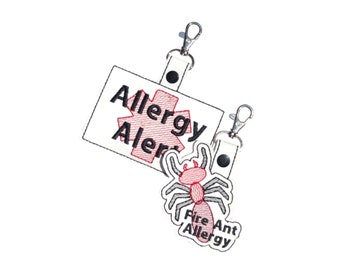 May include: Two white allergy alert tags with metal clasps. One tag has the words "Allergy Alert" with a red medical symbol. The other tag has a red and gray fire ant design with the words "Fire Ant Allergy".